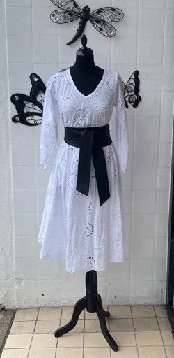 A white eyelet long-sleeve midi dress displayed on a black mannequin stand, featuring a black leather sash tied at the waist, set against a white brick wall with butterfly and dragonfly wall decorations.